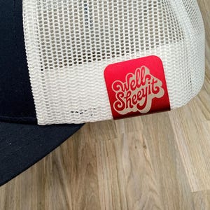 May include: A navy blue and white trucker hat featuring a white mesh back panel and a solid navy blue front panel. A rectangular red metal tag is attached to the side of the mesh, displaying the white text Well Sheeyit in a stylized font.