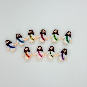 May include: A collection of small figurines depicting Jesus, each with a different colored sash. The figurines are wearing white robes and have brown hair and beards. The sashes have the text "Jesus Loves You".