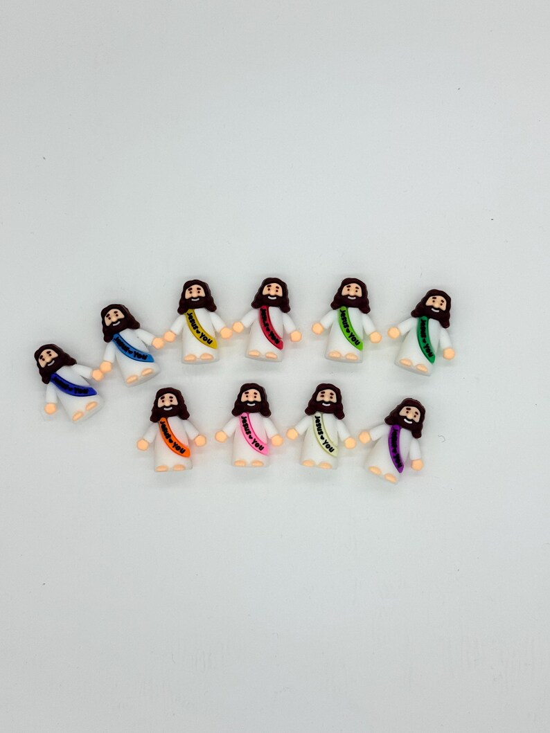 May include: A collection of small figurines depicting Jesus, each with a different colored sash. The figurines are wearing white robes and have brown hair and beards. The sashes have the text "Jesus Loves You".