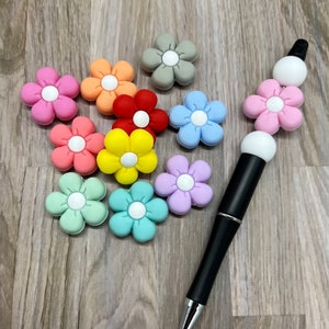 May include: A collection of colorful flower-shaped charms with white centers, arranged on a wooden surface. A black pen with white flower-shaped beads is also included.