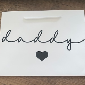 Puede incluir: Bolsa de regalo blanca con una inscripción en cursiva negra que dice "papá" y un corazón negro.