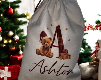 May include: A light gray Christmas gift bag with a drawstring closure. The bag features a teddy bear wearing a Santa hat, the letter "A" in a plaid pattern, and the name "Ashton" in a cursive font. The bag is decorated with small gold stars.