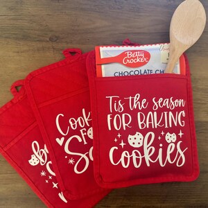 May include: Three red oven mitts with white text. The top mitt says "Tis the season for baking cookies" with two cookie images. The middle mitt says "Cook for Santa" with a cookie image. The bottom mitt says "Bake for Santa" with a cookie image.