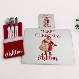 May include: A festive Christmas dining set. It includes a red and white cutlery holder, a placemat, and a coaster. The placemat and coaster feature a Santa Claus illustration, the text "Merry Christmas" and the name "Ashton".