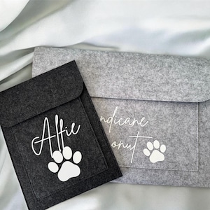 May include: Two felt pet accessory pouches. One is dark grey with the name "Alfie" and a paw print in white. The other is light grey with text and a paw print. The pouches are on a light grey fabric.