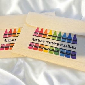 May include: Two cream-coloured felt pouches with a flap closure. Each pouch features a colourful crayon design and the text "Ashton's nursery creations" in black script. The crayons are arranged in a rainbow pattern.