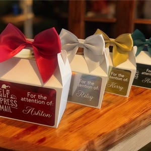 May include: Four white gift boxes with bows in red, silver, gold, and green. Each box has a label with a name and the text "Elf Express Mail". The boxes are on a wooden surface.