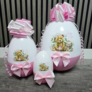 May include: A set of three white and pink Easter egg-shaped containers, each featuring a teddy bear illustration and the words "Happy Easter." They are decorated with pink bows and large pink and white ribbons. The eggs are of different sizes.