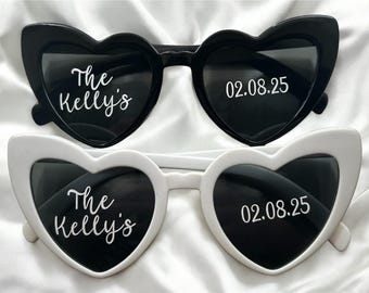 May include: Two pairs of heart-shaped sunglasses, one black and one white. The black sunglasses have the text "The Kelly's" and "02.08.25" in white. The white sunglasses also have the same text in white. The sunglasses are on a white background.