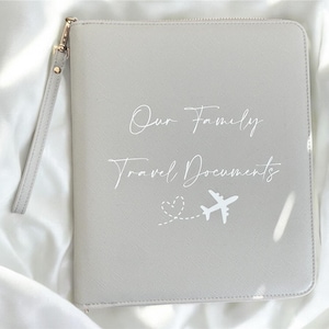 May include: A pale grey travel document wallet with a wrist strap and gold-coloured zip. The text "Our Family Travel Documents" is printed on the front, with a small airplane graphic.
