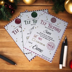 May include: Three "Elf Report" certificates on a wooden surface. Each certificate has a burgundy border, gold stars, and a wax seal. The text reads "North Pole Official Certificate" and "Nice". Pens and Christmas decorations are also visible.