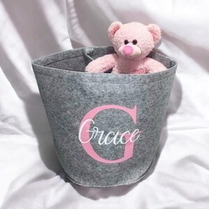 May include: A gray felt storage bin with a pink letter "G" and the name "Grace" written in white. A pink teddy bear is sitting inside the bin.