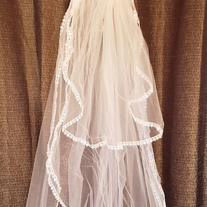 Puede incluir: Un velo de novia blanco con un borde festoneado y un delicado ribete de encaje. El velo está hecho de tul suave y tiene una sola capa.