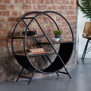 May include: A black metal circular shelf unit with four wooden shelves. The shelf unit has a unique design with a diagonal support structure. The shelf unit is standing on a grey floor in front of a brick wall.