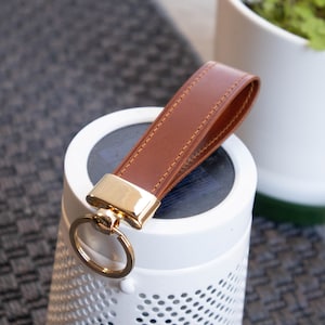 May include: A brown leather keychain with a gold metal ring and a stitched design. The keychain is attached to a white speaker with a perforated design.