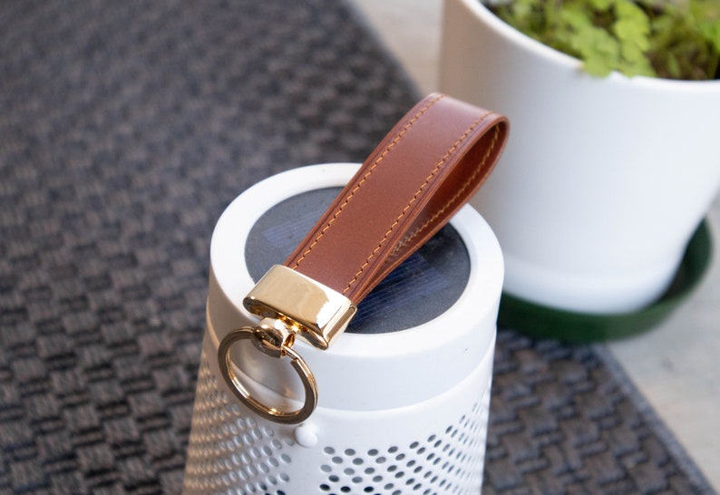 May include: A brown leather keychain with a gold metal ring and a stitched design. The keychain is attached to a white speaker with a perforated design.