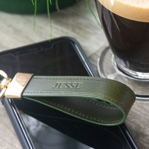 May include: A green leather keychain with a gold ring and the name "JESSE" embossed on it. The keychain is lying on top of a black smartphone.