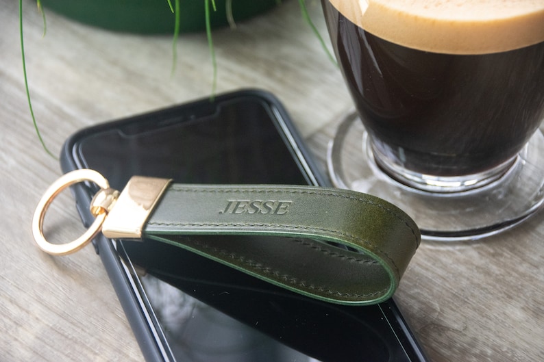 May include: A green leather keychain with a gold ring and the name "JESSE" embossed on it. The keychain is lying on top of a black smartphone.