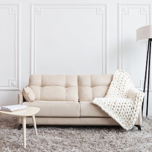 May include: A beige sofa with a white knitted throw blanket, a small wooden table with books on it, and a floor lamp with a white shade in a white room with decorative molding.