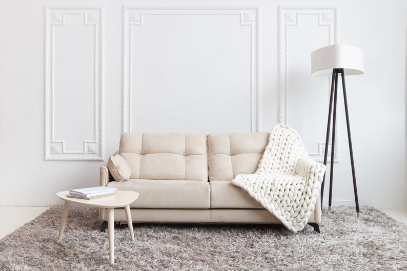 May include: A beige sofa with a white knitted throw blanket, a small wooden table with books on it, and a floor lamp with a white shade in a white room with decorative molding.