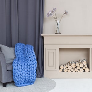 May include: A blue chunky knit throw blanket draped over a gray armchair with a gray pillow. The armchair is in front of a white fireplace with a mantel and a vase of dried flowers.