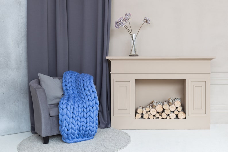 May include: A blue chunky knit throw blanket draped over a gray armchair with a gray pillow. The armchair is in front of a white fireplace with a mantel and a vase of dried flowers.