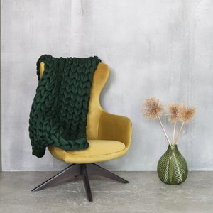 May include: A green knitted throw draped over a yellow velvet armchair with a black wooden base. A green vase with dried flowers sits on the floor.