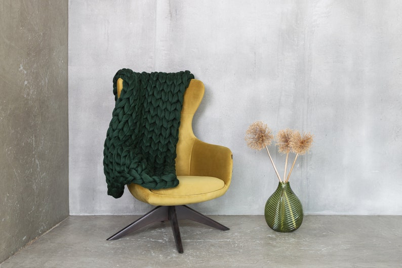 May include: A green knitted throw draped over a yellow velvet armchair with a black wooden base. A green vase with dried flowers sits on the floor.