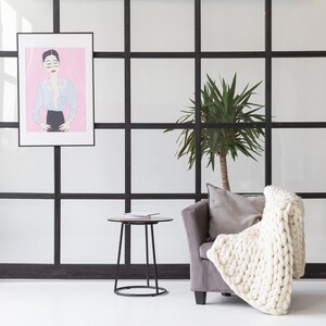 May include: A white knitted throw blanket draped over a gray armchair with a black frame. A potted green plant is in the background, and a black side table with a book on top is in the foreground.