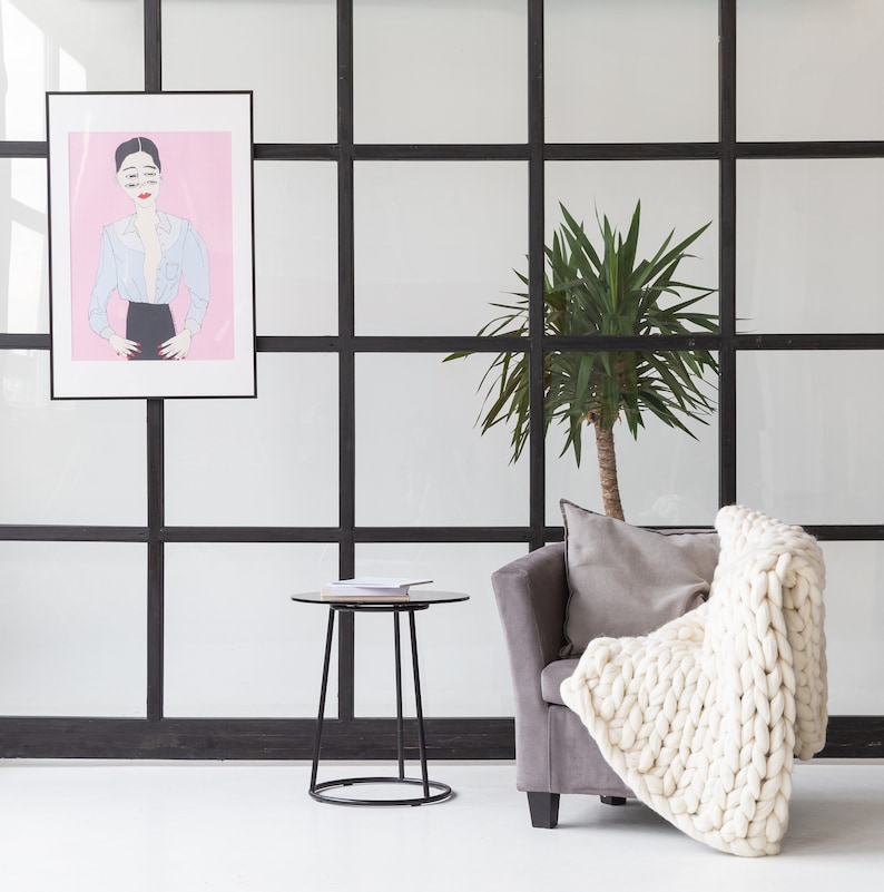 May include: A white knitted throw blanket draped over a gray armchair with a black frame. A potted green plant is in the background, and a black side table with a book on top is in the foreground.