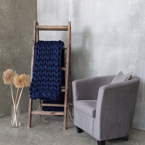 May include: A gray armchair with a pillow sits in front of a wooden ladder with a navy blue chunky knit blanket draped over it. The ladder and chair are in front of a gray concrete wall.