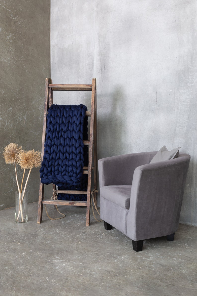 May include: A gray armchair with a pillow sits in front of a wooden ladder with a navy blue chunky knit blanket draped over it. The ladder and chair are in front of a gray concrete wall.