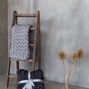 May include: A gray chunky knit throw blanket draped over a wooden ladder with a black chunky knit pillow tied with a silver ribbon in front of a gray concrete wall.
