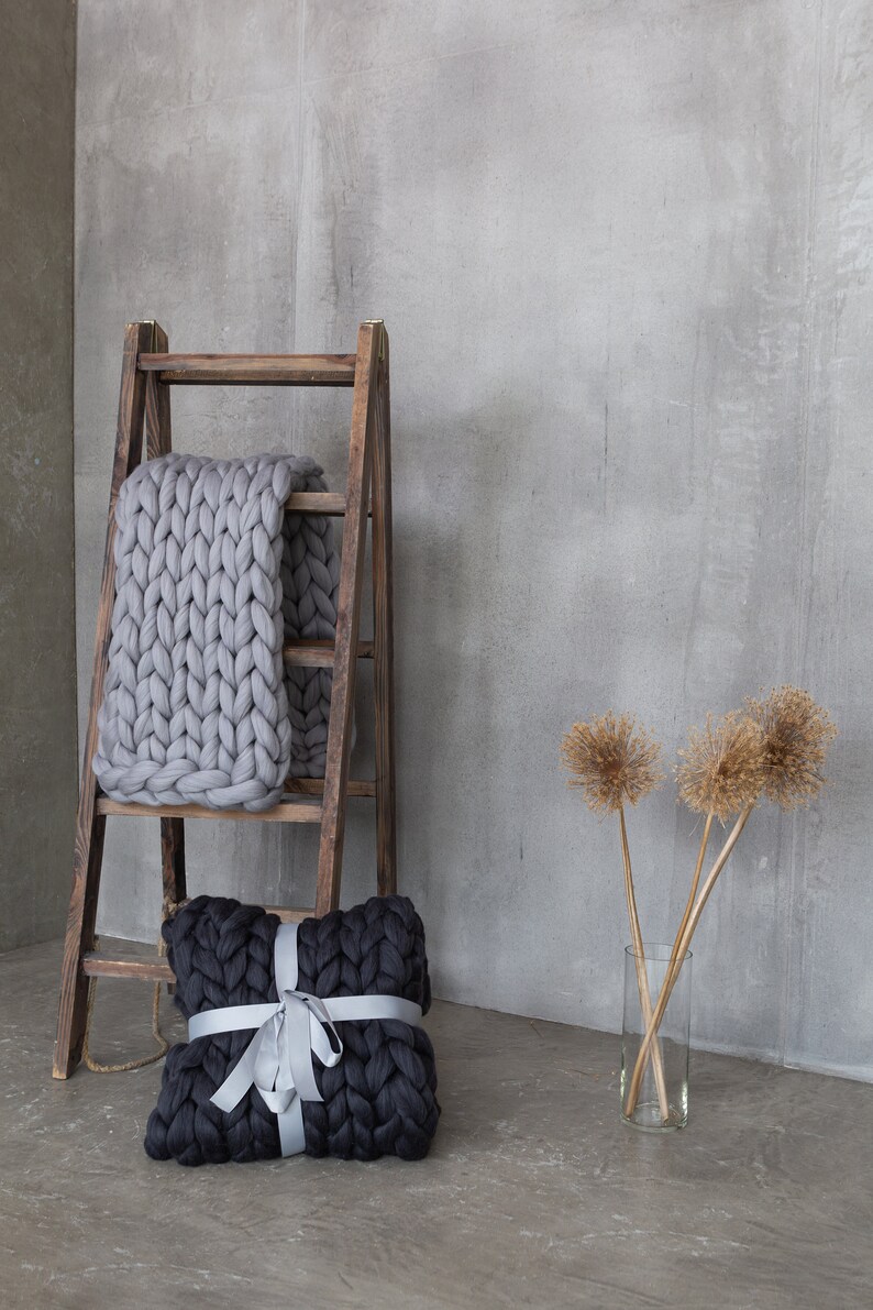May include: A gray chunky knit throw blanket draped over a wooden ladder with a black chunky knit pillow tied with a silver ribbon in front of a gray concrete wall.