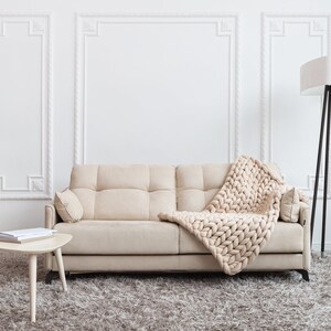 May include: A beige sofa with a chunky knit throw blanket draped over it. The sofa is in a living room with a white wall, a white lamp, and a small wooden table with books on it.