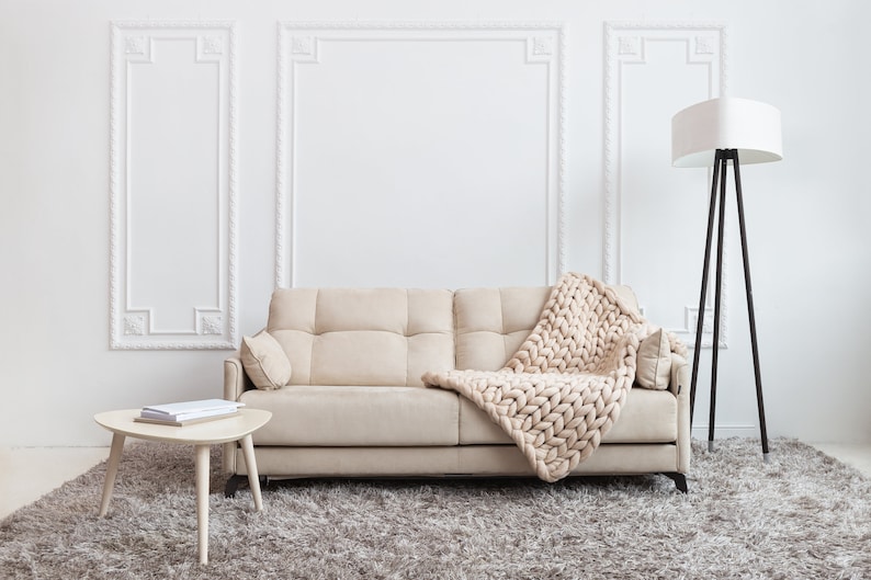 May include: A beige sofa with a chunky knit throw blanket draped over it. The sofa is in a living room with a white wall, a white lamp, and a small wooden table with books on it.