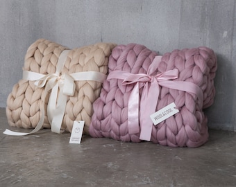 Puede incluir: Dos almohadas grandes de punto grueso, una en beige y otra en rosa polvoriento, atadas con una cinta de satén. Las almohadas están hechas de lana y tienen una textura tejida. Las almohadas están sobre una superficie de hormigón gris.