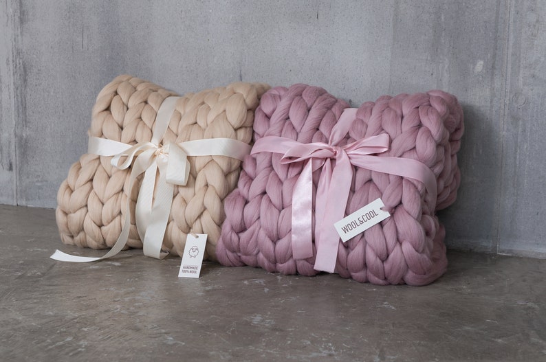 May include: Two large, chunky knit throw pillows, one in beige and one in dusty rose, tied with satin ribbon. The pillows are made of wool and have a woven texture. The pillows are sitting on a gray concrete surface.