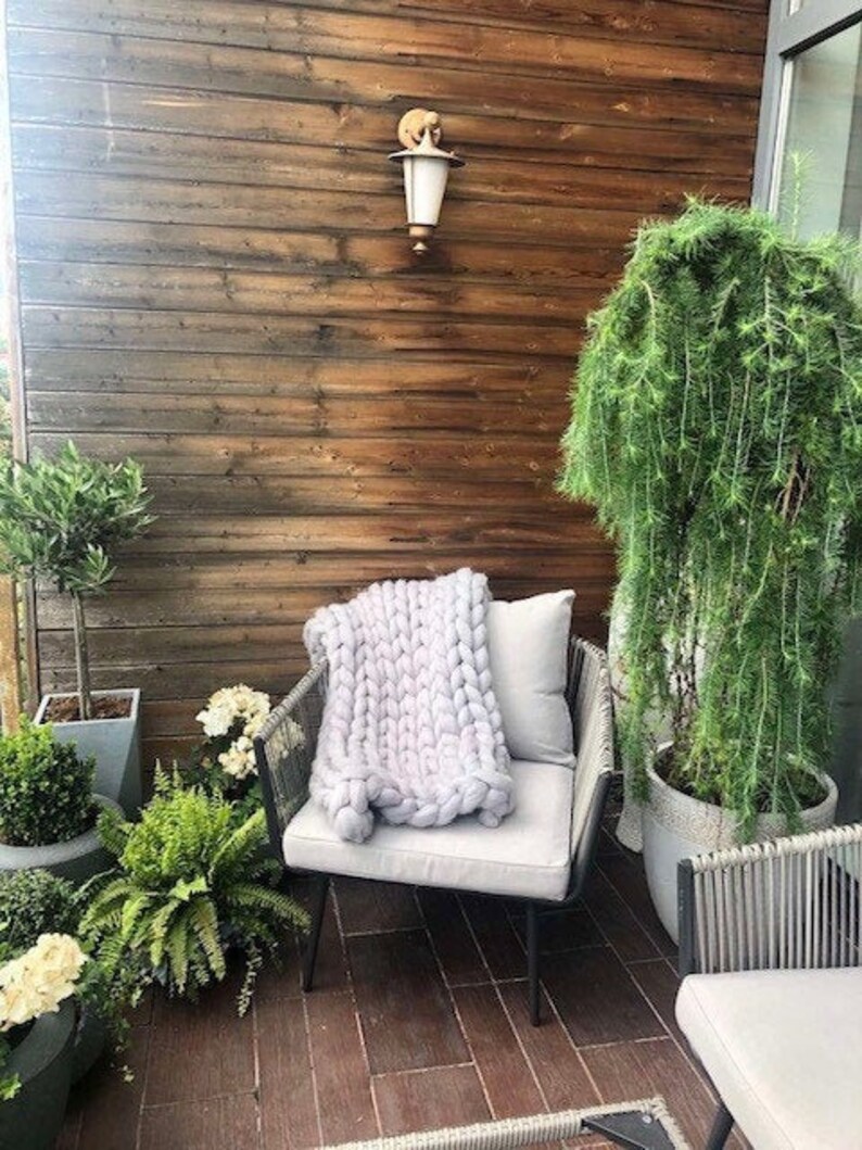 May include: A light gray chunky knit throw blanket draped over a black metal chair with a white cushion. The chair is on a patio with a wooden wall and potted plants.