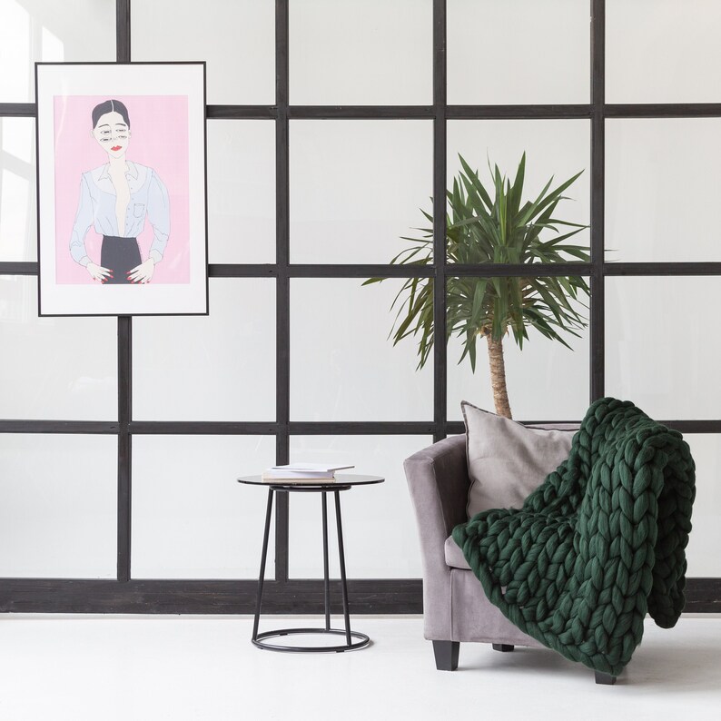 May include: A green knitted throw blanket draped over a gray armchair with a black metal side table and a potted plant in the background.