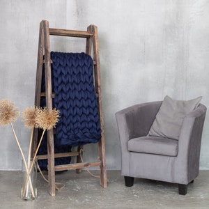 May include: A wooden ladder with a navy blue knitted throw blanket draped over it. A gray armchair with a gray pillow sits in front of the ladder. Dried flowers are in a glass vase on the floor.
