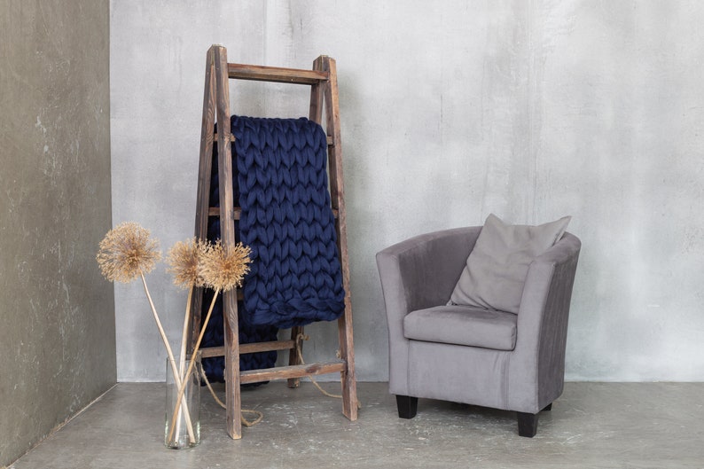 May include: A wooden ladder with a navy blue knitted throw blanket draped over it. A gray armchair with a gray pillow sits in front of the ladder. Dried flowers are in a glass vase on the floor.