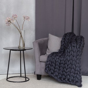 May include: A gray armchair with a chunky knit throw blanket draped over it. The chair is in front of a gray curtain and a small black side table with a vase of dried flowers.