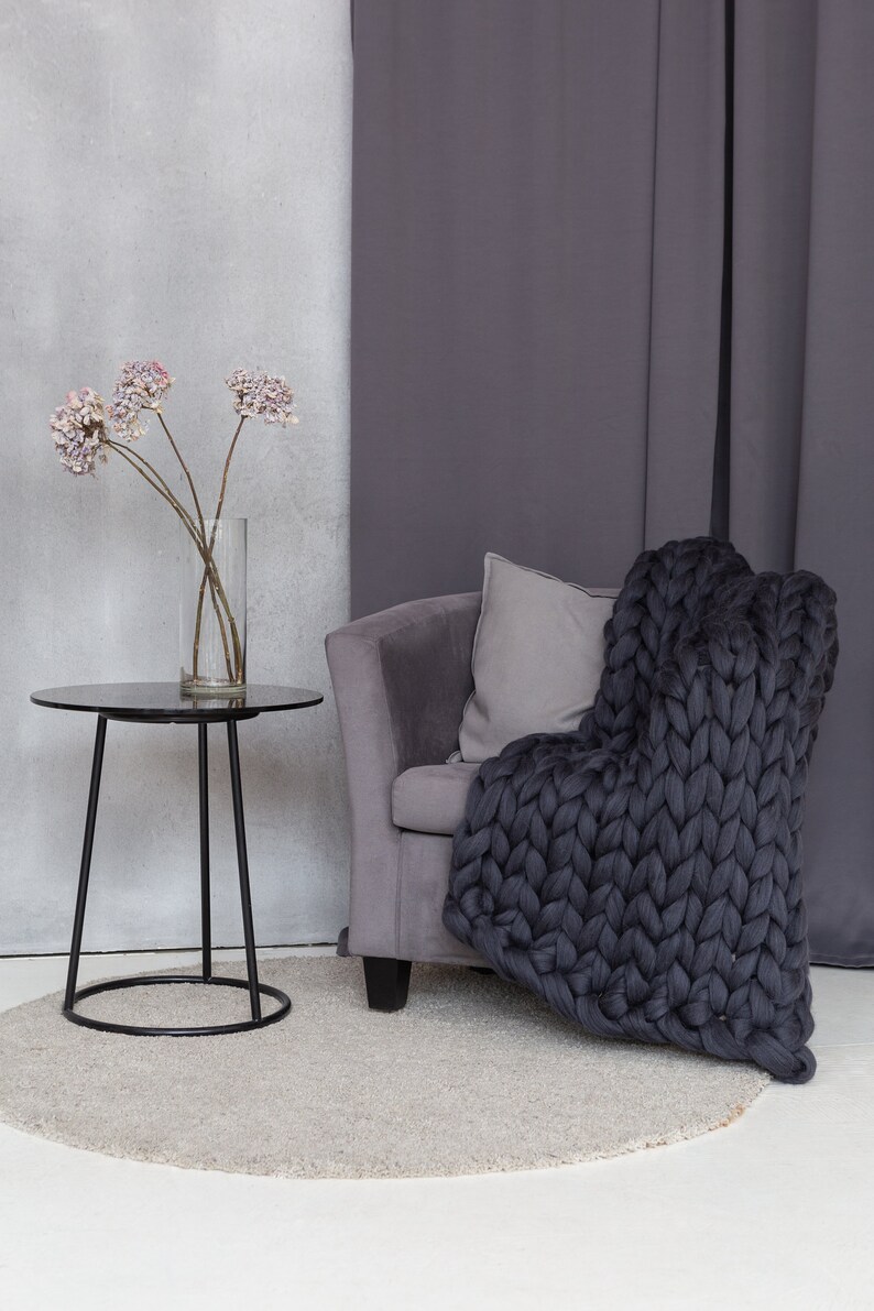 May include: A gray armchair with a chunky knit throw blanket draped over it. The chair is in front of a gray curtain and a small black side table with a vase of dried flowers.