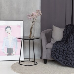 May include: A black side table with a round marble top sits in a living room with a gray armchair and a chunky knit throw blanket. A vase with dried flowers sits on the table.