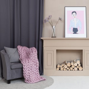 May include: A gray armchair with a pink knitted throw blanket and a gray pillow sits in front of a beige fireplace with a framed illustration of a woman in a blue shirt and black skirt. The fireplace has a stack of logs in the hearth.