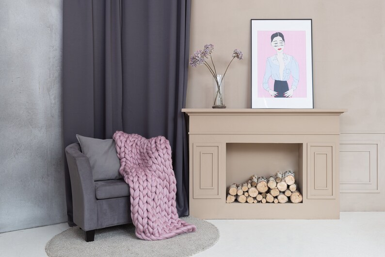 May include: A gray armchair with a pink knitted throw blanket and a gray pillow sits in front of a beige fireplace with a framed illustration of a woman in a blue shirt and black skirt. The fireplace has a stack of logs in the hearth.