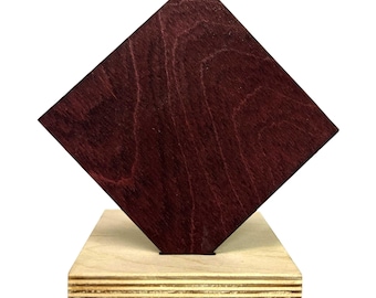 May include: A wooden sculpture featuring a dark burgundy diamond shape mounted on a light-colored rectangular base. The diamond is angled, showcasing the wood grain. The base is made of layered wood, creating a contrast in texture and color.
