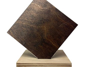 May include: A wooden sculpture featuring a dark brown, square-shaped piece of wood set at an angle on a light brown, rectangular base. The wood grain is visible on both pieces, creating a natural, minimalist aesthetic.