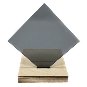 May include: A dark gray, square-shaped piece of material is displayed on a light-colored wooden base. The material is angled, creating a diamond shape. The wooden base is rectangular and appears to be made of layered wood.